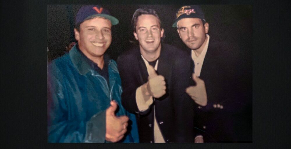 "Three men posing together, all smiling and giving thumbs up; two wear baseball caps and casual jackets, while one in the middle is in a dark blazer. The background is dark, suggesting an indoor or nighttime setting."